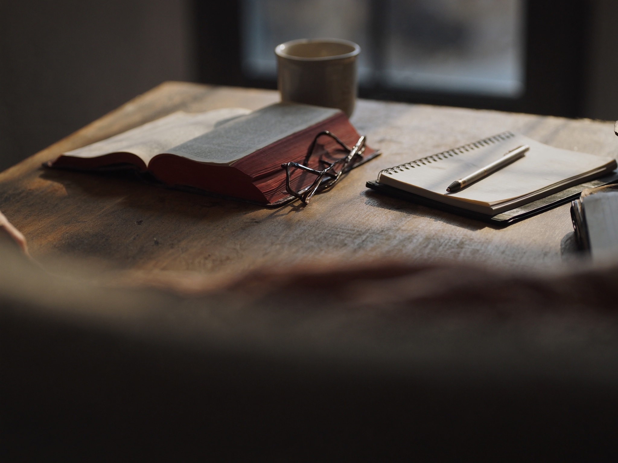 An open Bible on a wooden table beside a notebook, pen and reading glasses in morning light